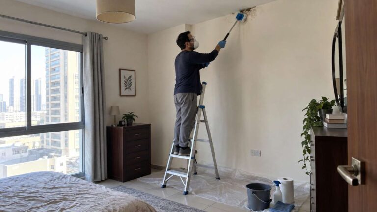 Homeowner safely cleaning a small mold patch high on a ceiling wall using a stable ladder and protective equipment