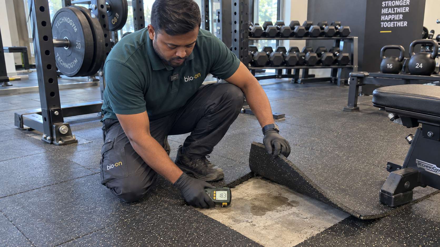 Bio-On technician checking damp substrate beneath lifted rubber flooring in a commercial gym.