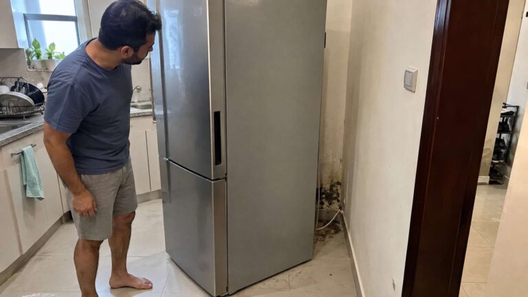 Refrigerator pulled away from a kitchen wall revealing localized mold and moisture damage behind it.