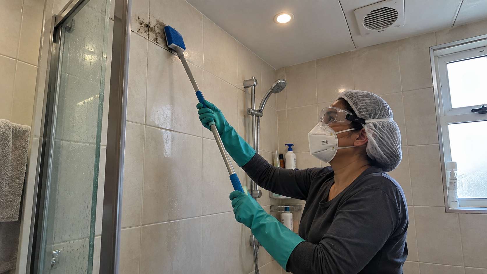 Homeowner wearing N95 respirator and sealed goggles while cleaning mold above shoulder height in a shower