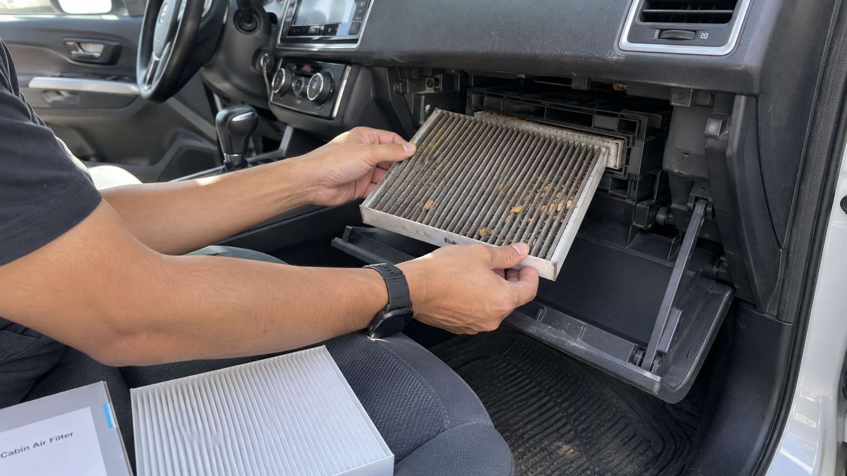 Dirty cabin air filter being removed from behind the glovebox beside a clean replacement filter.
