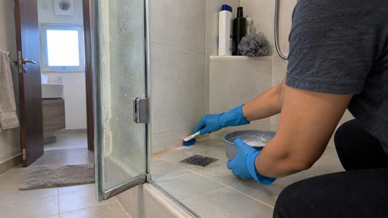 Homeowner scrubbing pink slimy biofilm from a shower grout and silicone corner.