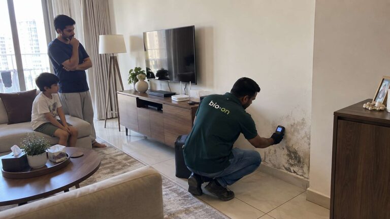 Bio-On technician checking a damp mold-affected wall in a UAE family living room while an adult and child observe from farther away.