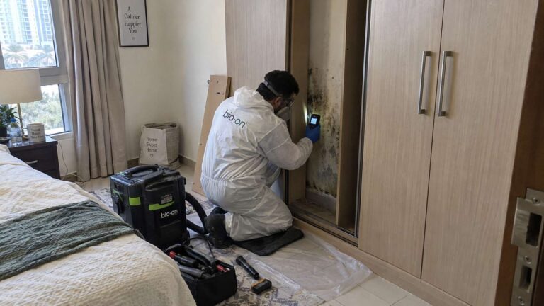 Mold remediation technician inspecting a damp narrow space behind built-in furniture.