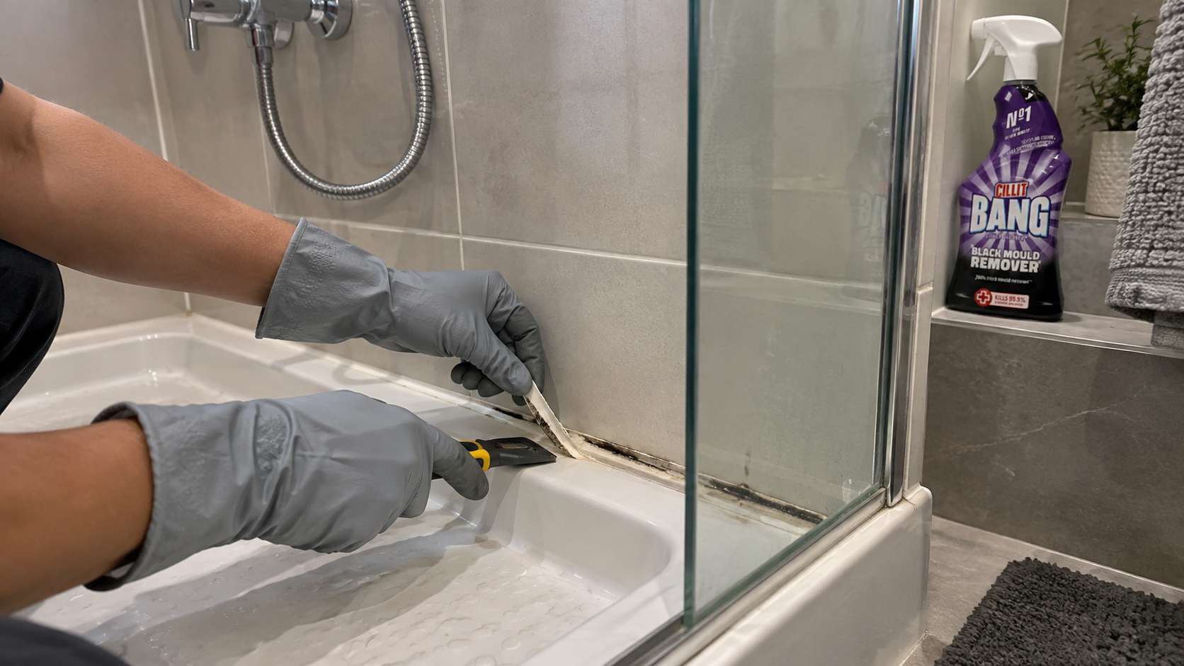 Worker removing deeply mould-stained silicone sealant from a shower edge.